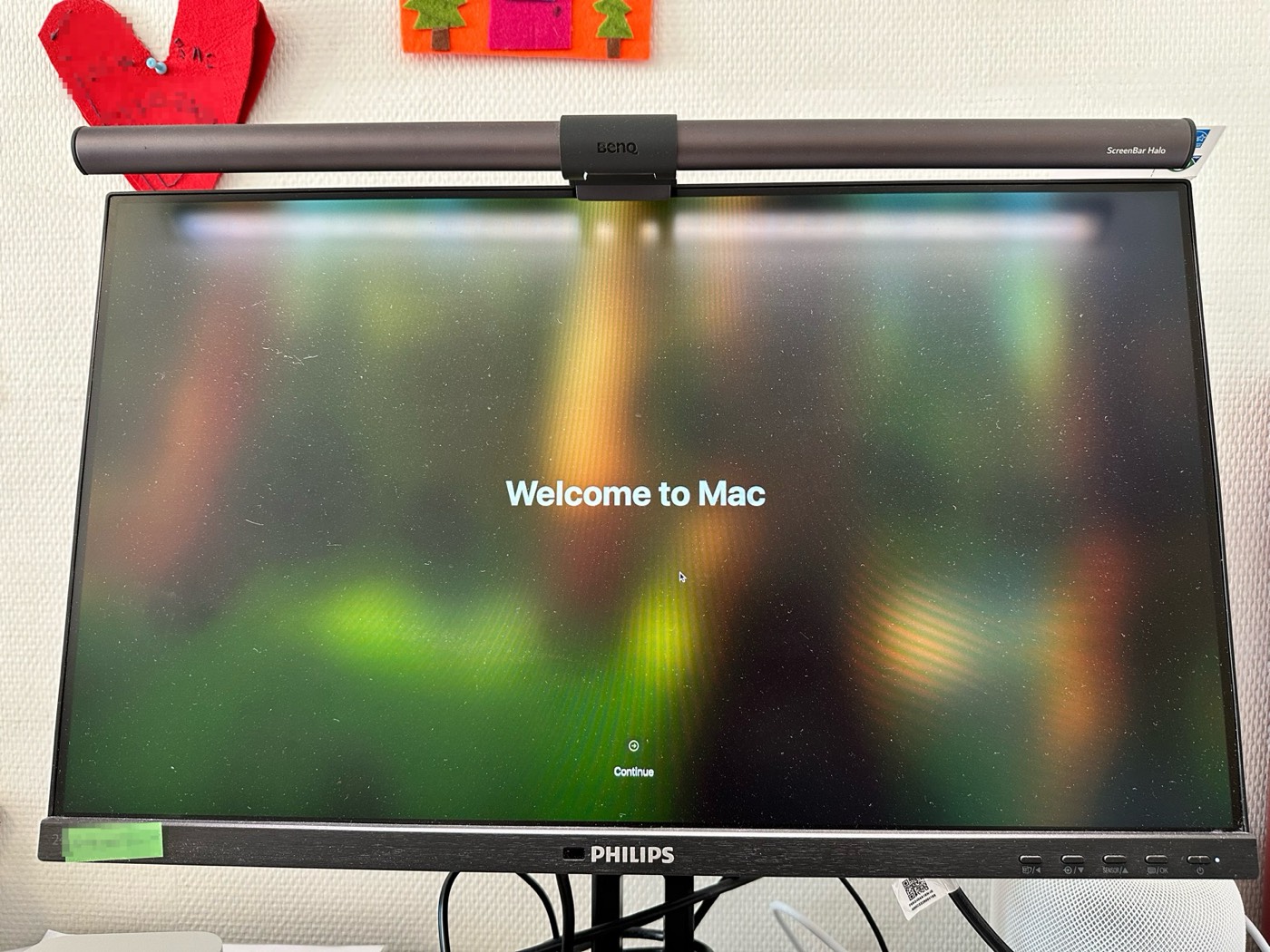 A Philips monitor on a desk showing the macOS setup assistant's Welcome to Mac screen, lit by a BenQ screen bar