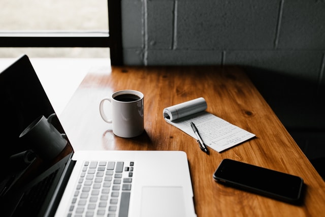 An open laptop on a wooden desk beside a mug of black coffee, a notepad with a pen, and a phone